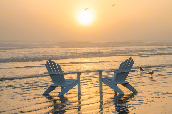 A photo of a pair of Adirondack Chairs on the beach at sunrise with sea fog 