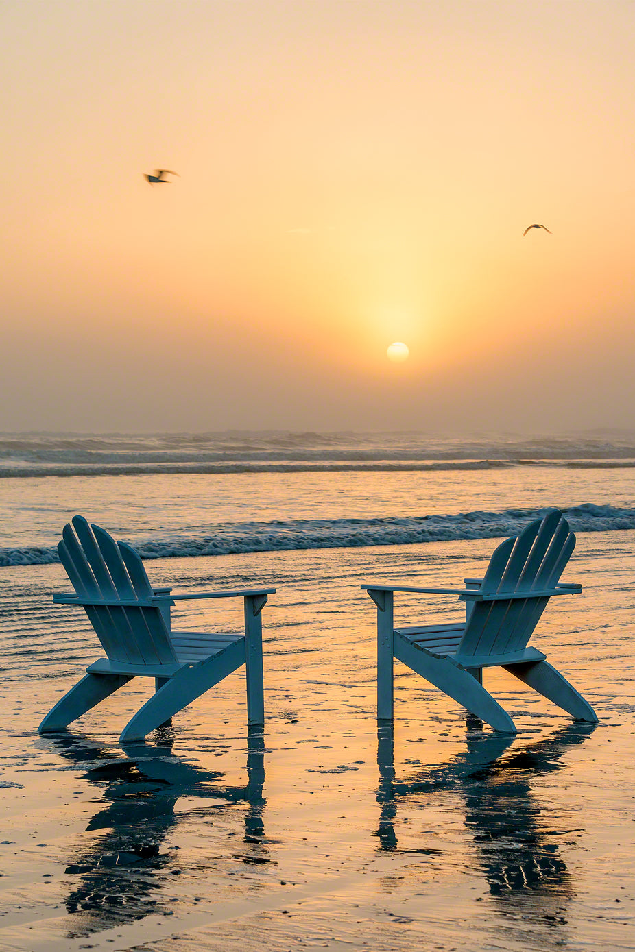 A photo of a pair of Adirondack Chairs on the beach at sunrise with sea fog